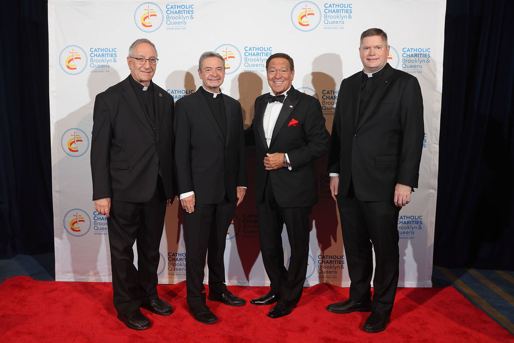2023 Bishop's Humanitarian Award Dinner with Joe Piscopo Group photo of 4 smiling men dressed in black on a red carpet in front of a step and repeat.